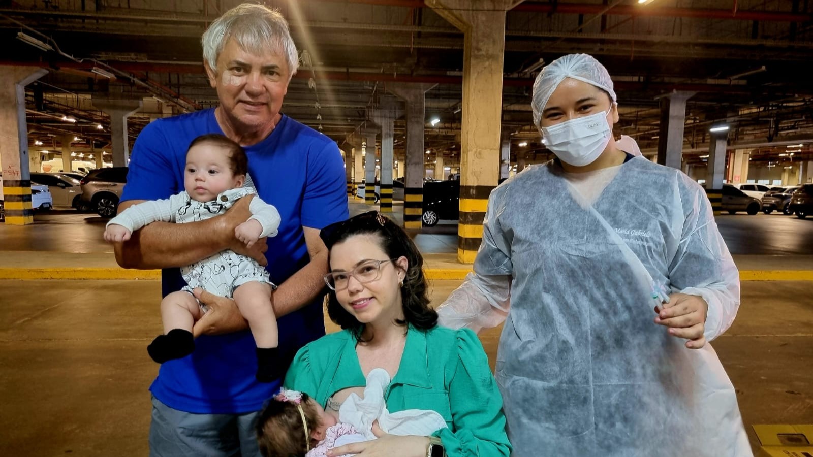 Camila Carvalho segura uma das filhas gêmeas, ao lado dela, um senhor segura a outra bebê e uma enfermeira está ao lado deles